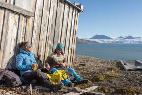 Bilde av to damer som sitter i solveggen med natur og bre i bakgrunnen på Svalbard.